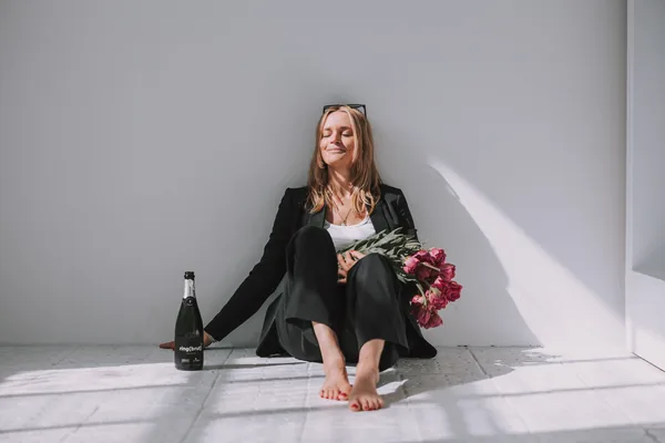 Frau sitzt mit Blumen im Sonnenlicht an einer weißen Wand